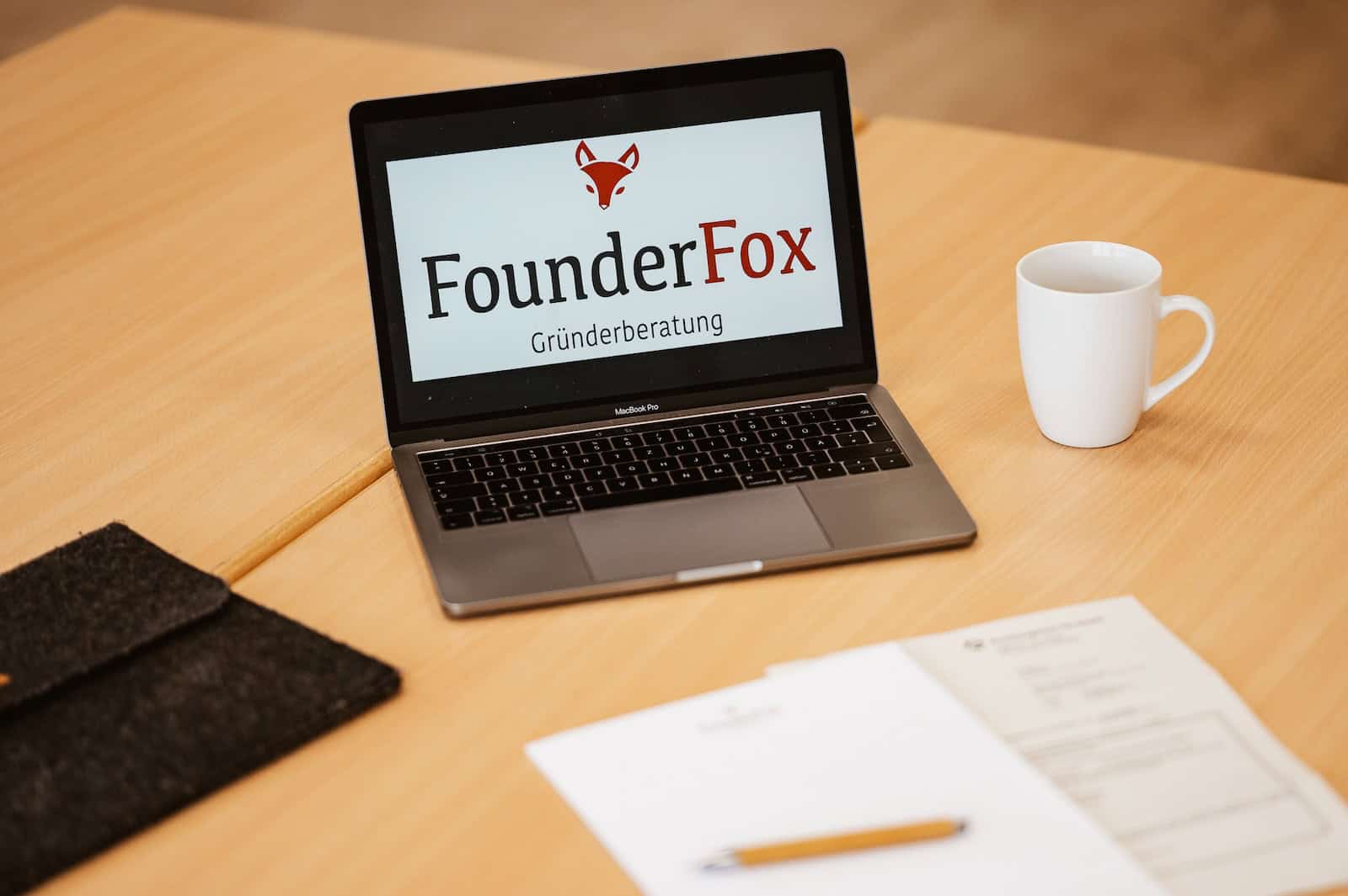 Offenes Notebook mit dem Logo von FounderFox Gründerberatung. Daneben ein Kaffeebecher, Formulare, ein Notizblock und Stift.