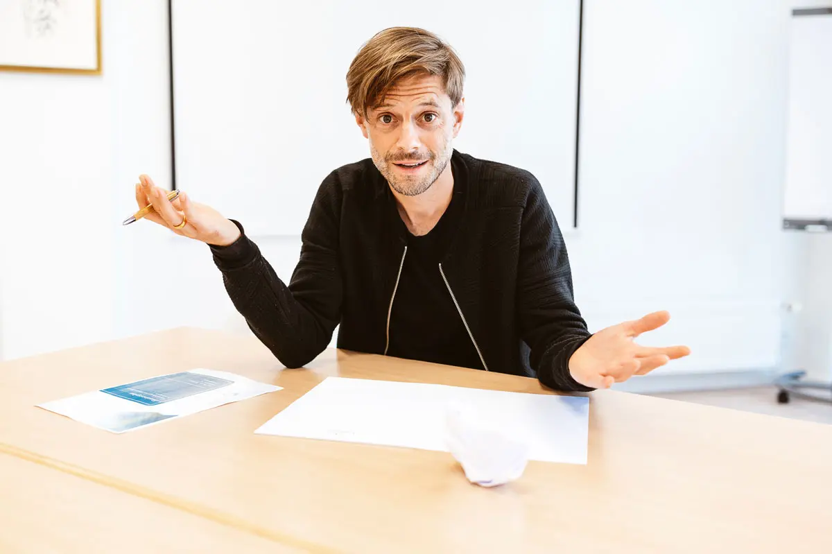 Mann mit fragender Geste am Schreibtisch mit Unterlagen