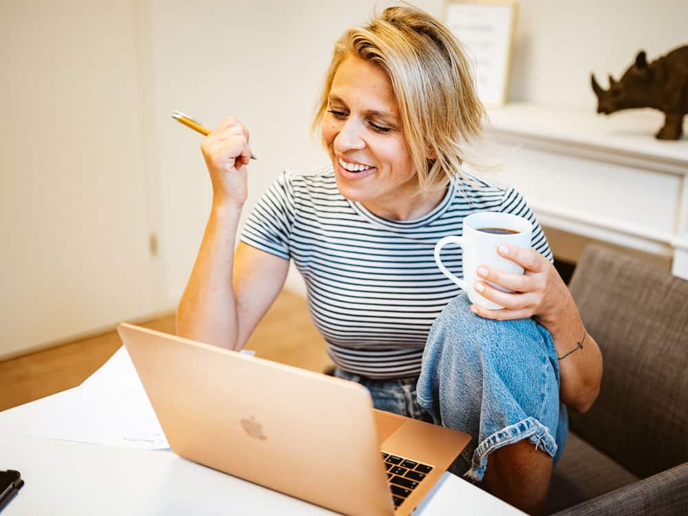 Eine junge Frau sitzt am Notebook mit einer Tasse Kaffee in der Hand und füllt gut gelaunt den Antrag auf ein AVGS-Gründercoaching aus