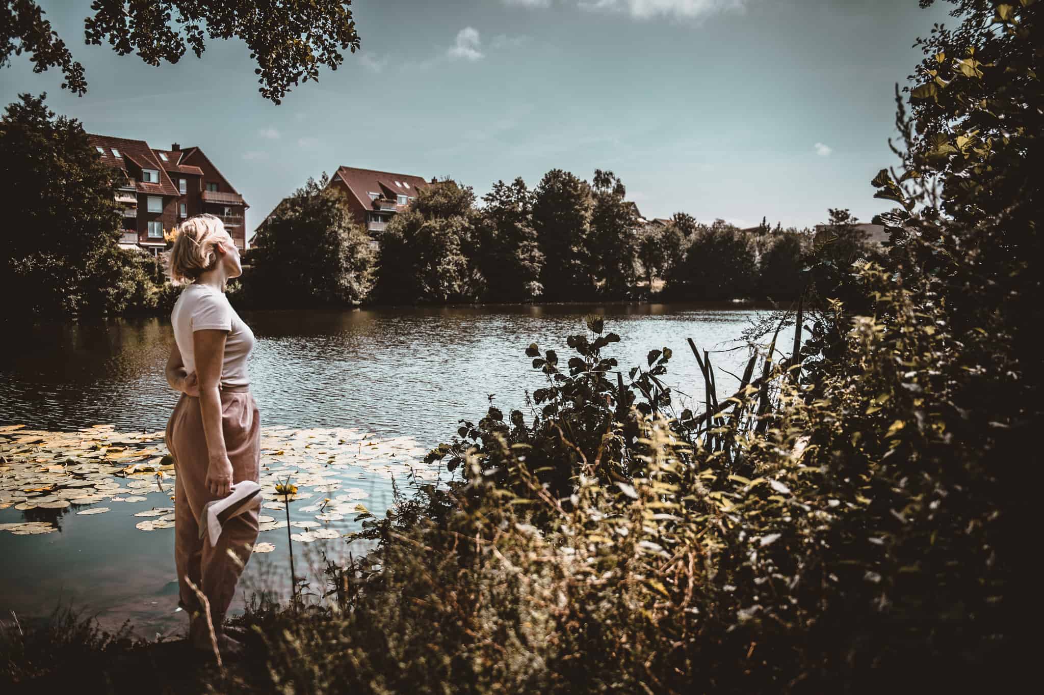 Eine Frau steht an einem ruhigen, von Grün umgebenen See und blickt auf das Wasser. Im Hintergrund sind Häuser zu sehen. Der Himmel ist teilweise bewölkt und Seerosen schwimmen friedlich auf der Oberfläche des Sees, ganz ähnlich wie AVGS Karrierecoaching einen durch Karrierereflexionen führt.